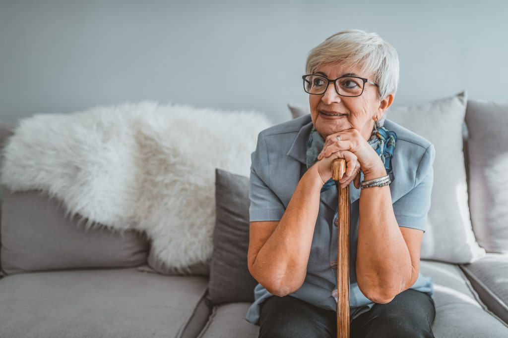Retired short hair old woman with her walking stick at home looking away during the day while sitting on sofa. Senior gray hair women supporting on a walking cane at home.