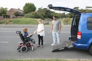 6 year old child with muscular dystrophy steering his electric wheelchair to gain access to family car, mother and father assisting and encouraging