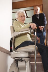 Smiling 93 year old senior on her stairlift in her home with another senior using a cane looking on.