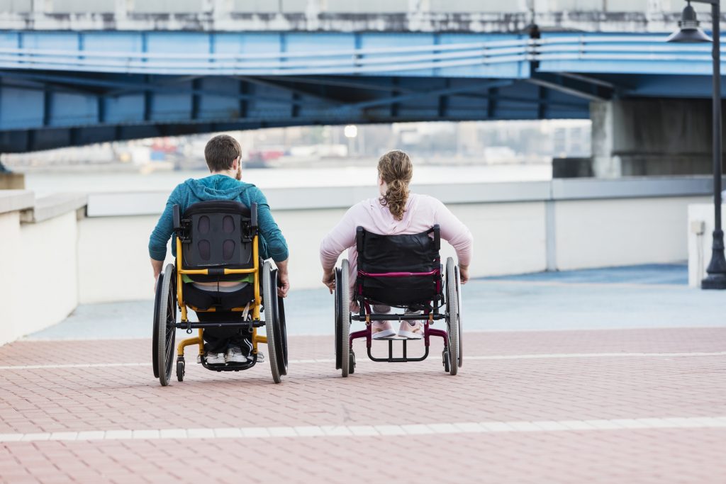 Rear view of a couple in wheelchairs side by side outdoors in the city, traveling down a pedestrian walkway. The young woman, in her 20s, has spina bifida and her mate, a mid adult man in his 30s, has cerebral palsy.