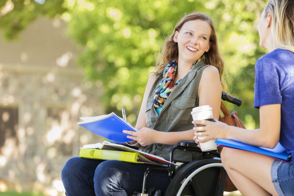 Pretty college girl in wheelchair talks with friend before class. She is holding a notebook and her friend is holding a coffee cup. She has long brown hair and is wearing a vest and a scarf.