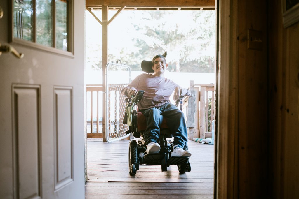 A cheerful young adult man with cerebral palsy drives up a wheelchair ramp to enter his home. Horizontal image with copy space.
